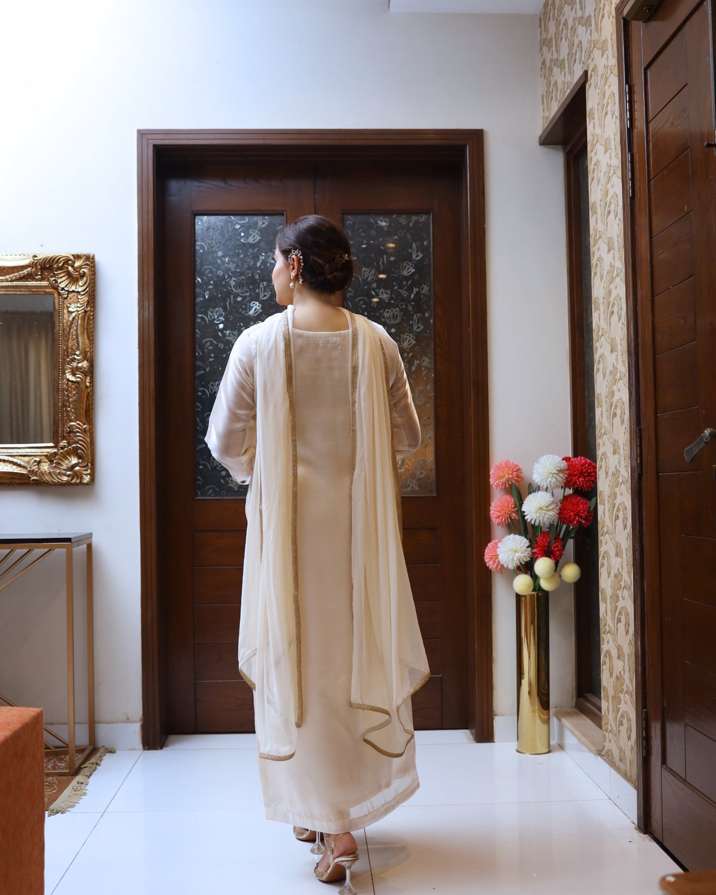 Woman in a beige outfit standing in a room with a decorative mirror and floral arrangement.