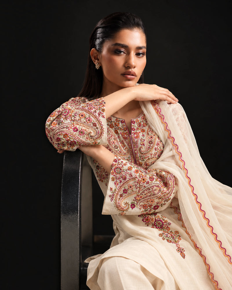 A woman seated on a black chair wearing an embellished cream-colored traditional outfit with floral and paisley embroidery in pink, red, and gold, against a dark studio background.