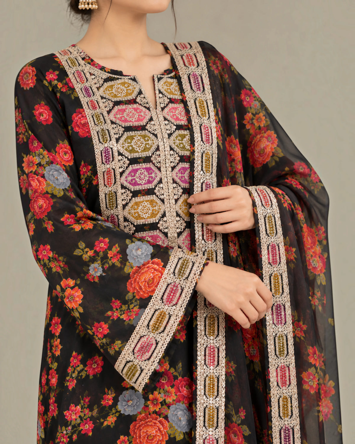 A close-up of a woman wearing a black floral salwar kameez suit with intricate multicolor embroidery and a matching dupatta, shown against a plain gray background.