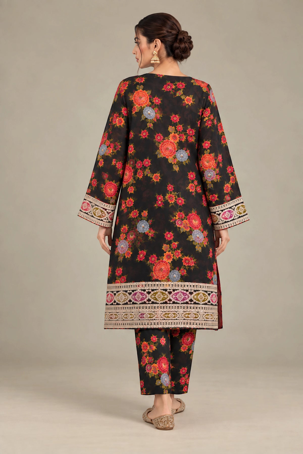 A woman shown from the back wearing a black three-piece traditional outfit with a vibrant multicolor floral print and ornate embroidered borders on the sleeves and hem, set against a plain beige studio background.