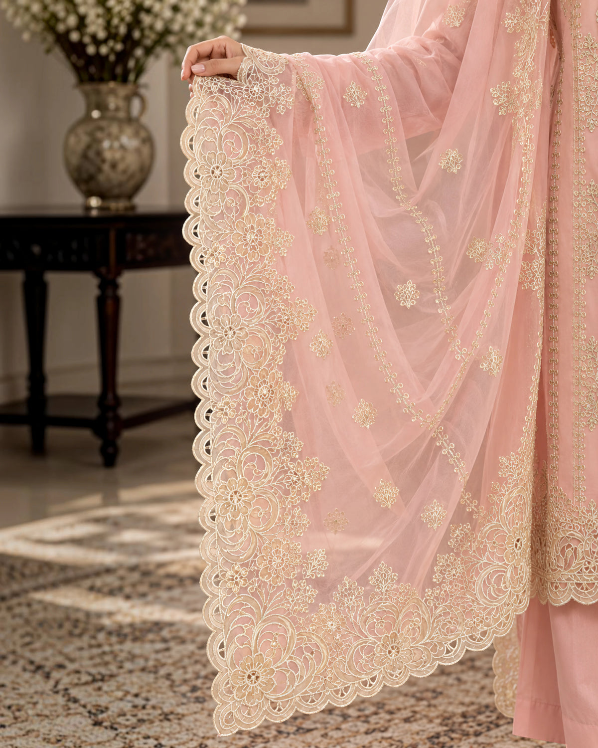 A close-up of a person holding up a light pink embroidered dupatta or scarf with ornate cream floral lace borders and mirror work, shown in an indoor room setting.