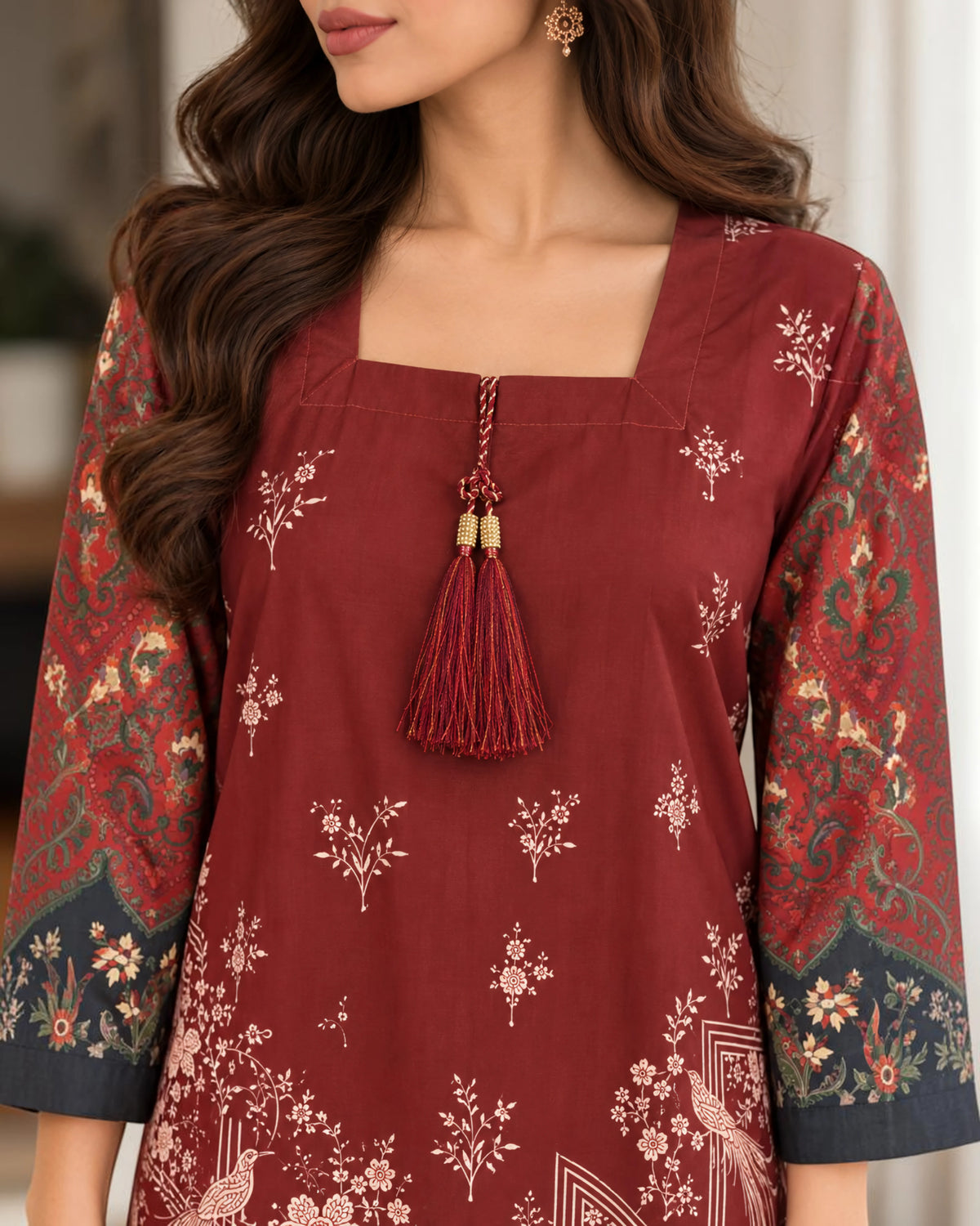 A woman wearing a deep red kurta with beige floral prints, featuring a square neckline, a decorative tassel at the center, and patterned sleeves with dark navy borders.