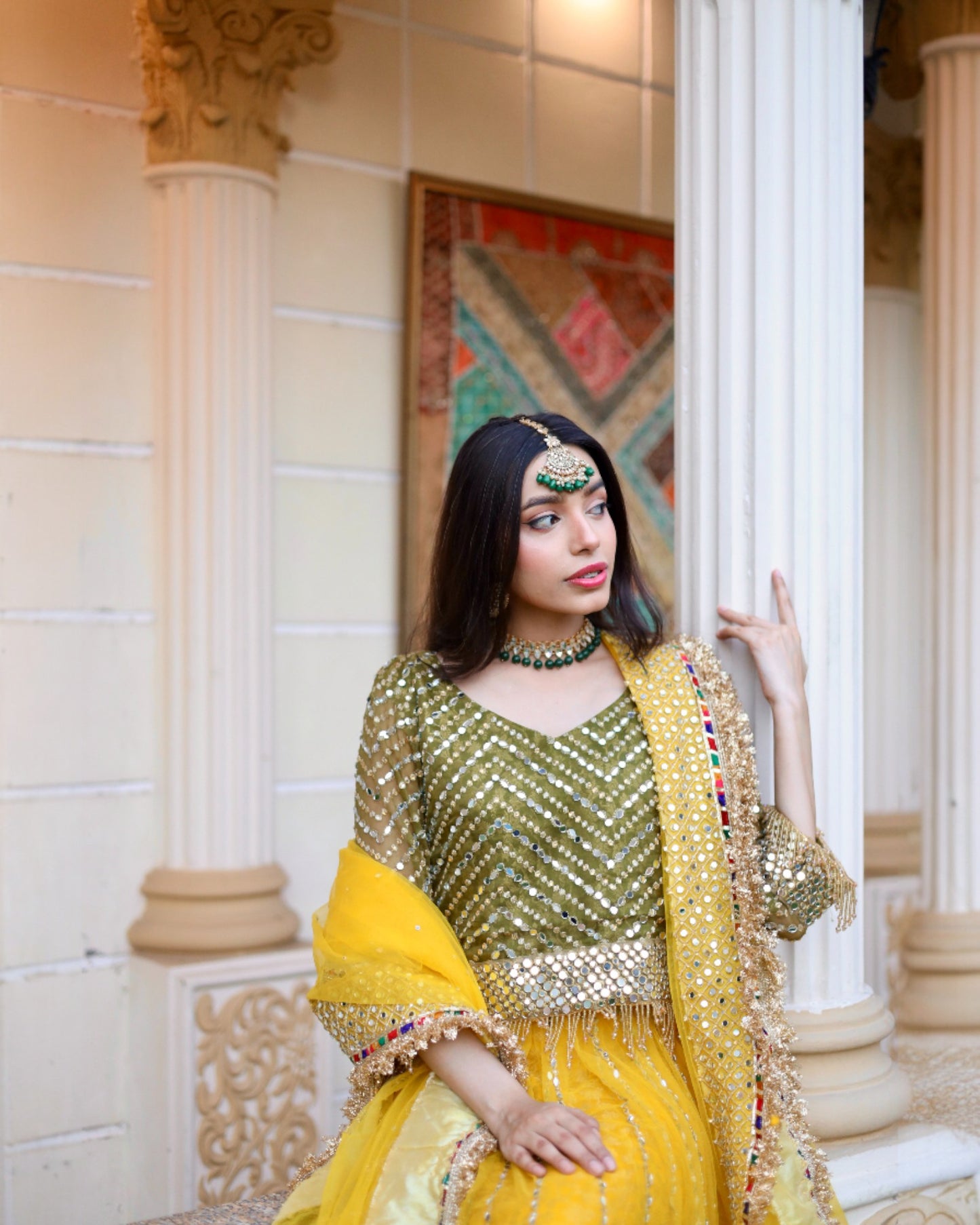 Woman in traditional yellow and green outfit with jewelry, standing indoors.