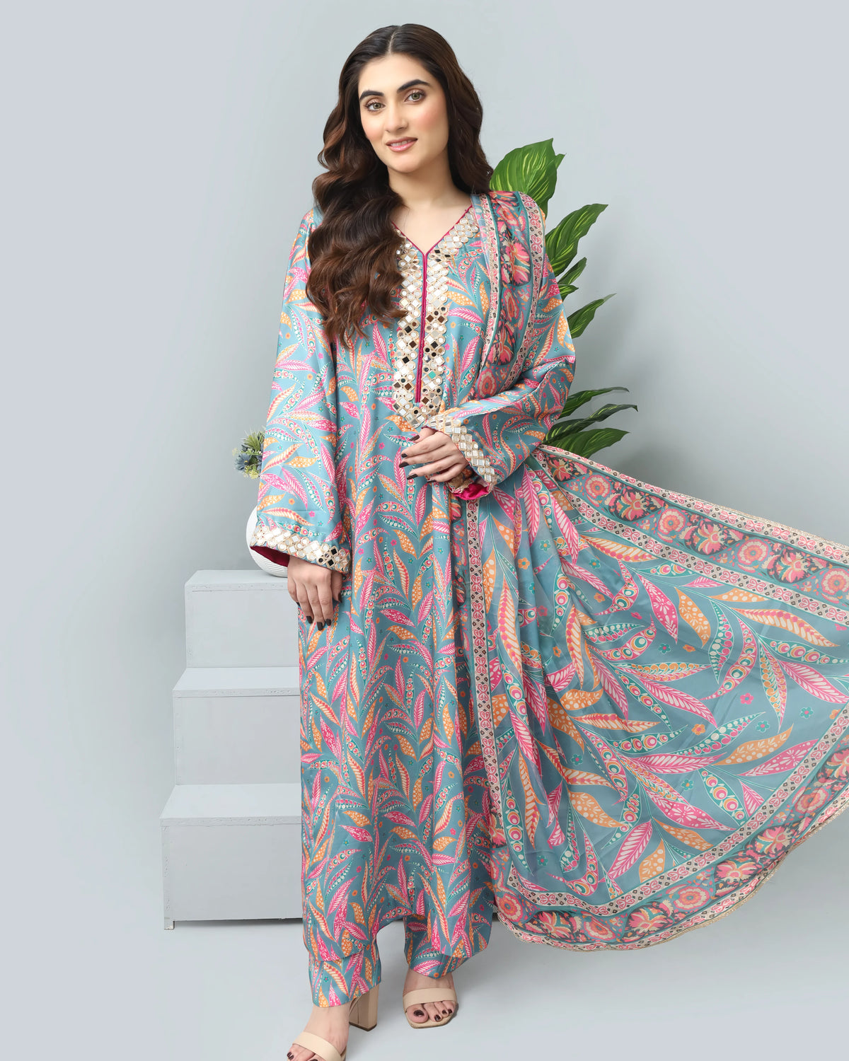 A woman models a three-piece printed ethnic outfit consisting of a long kurta, matching trousers, and a dupatta. The fabric features a vibrant multicolor pattern of pink, orange, and yellow leaves on a dusty teal-blue background.