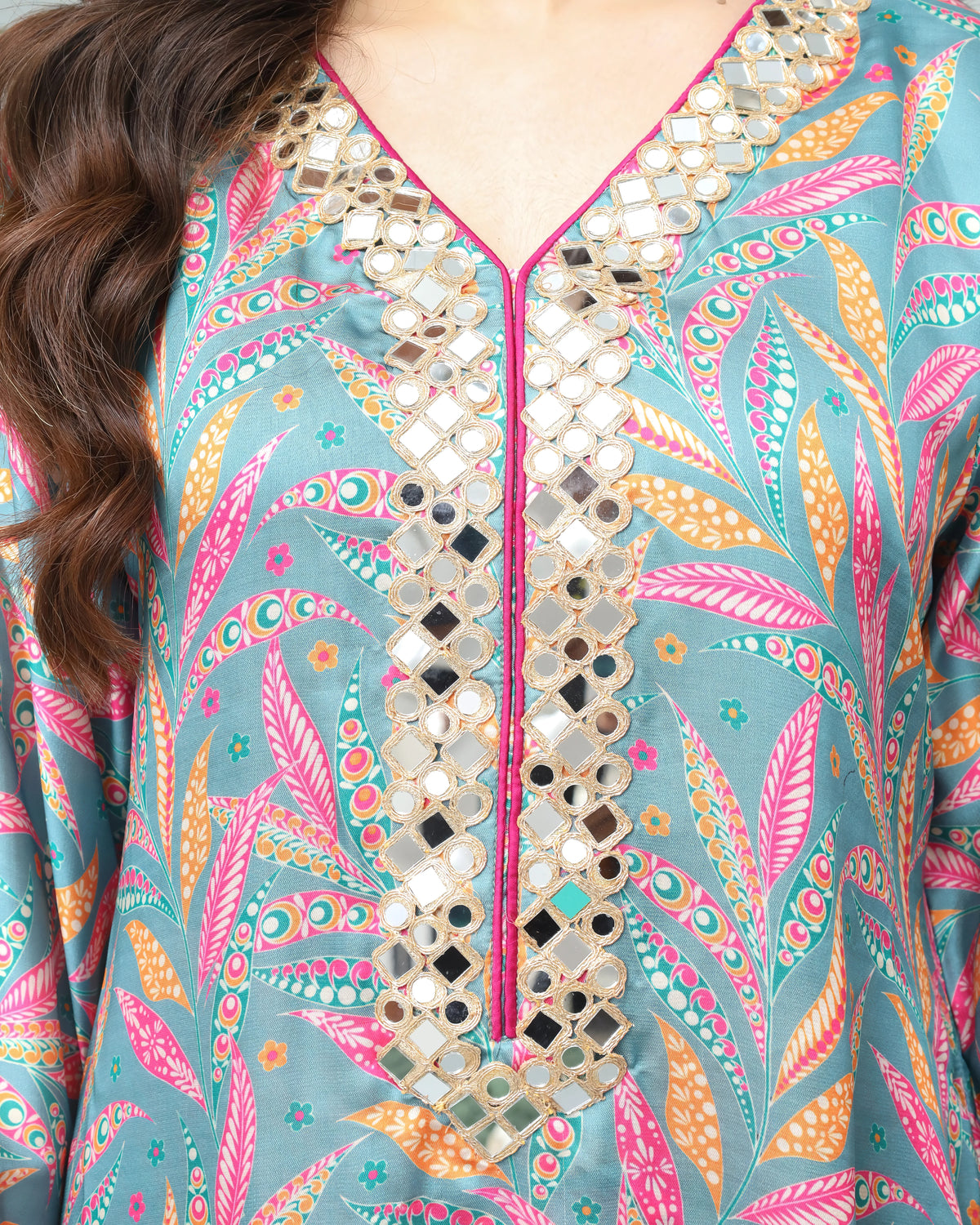A close-up of a women's long-sleeve tunic top featuring a teal blue base with a vibrant pink, orange, and yellow paisley pattern, detailed with a central mirror-work neckline and pink piping.