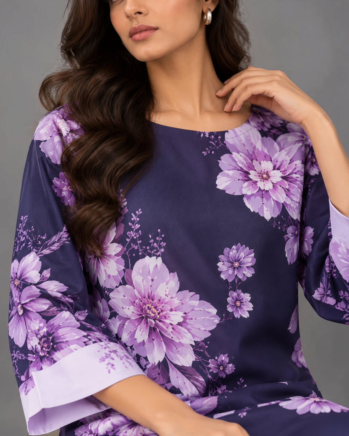 A woman wearing a purple floral print tunic with wide three-quarter sleeves and a boat neckline, shown against a plain gray studio background.