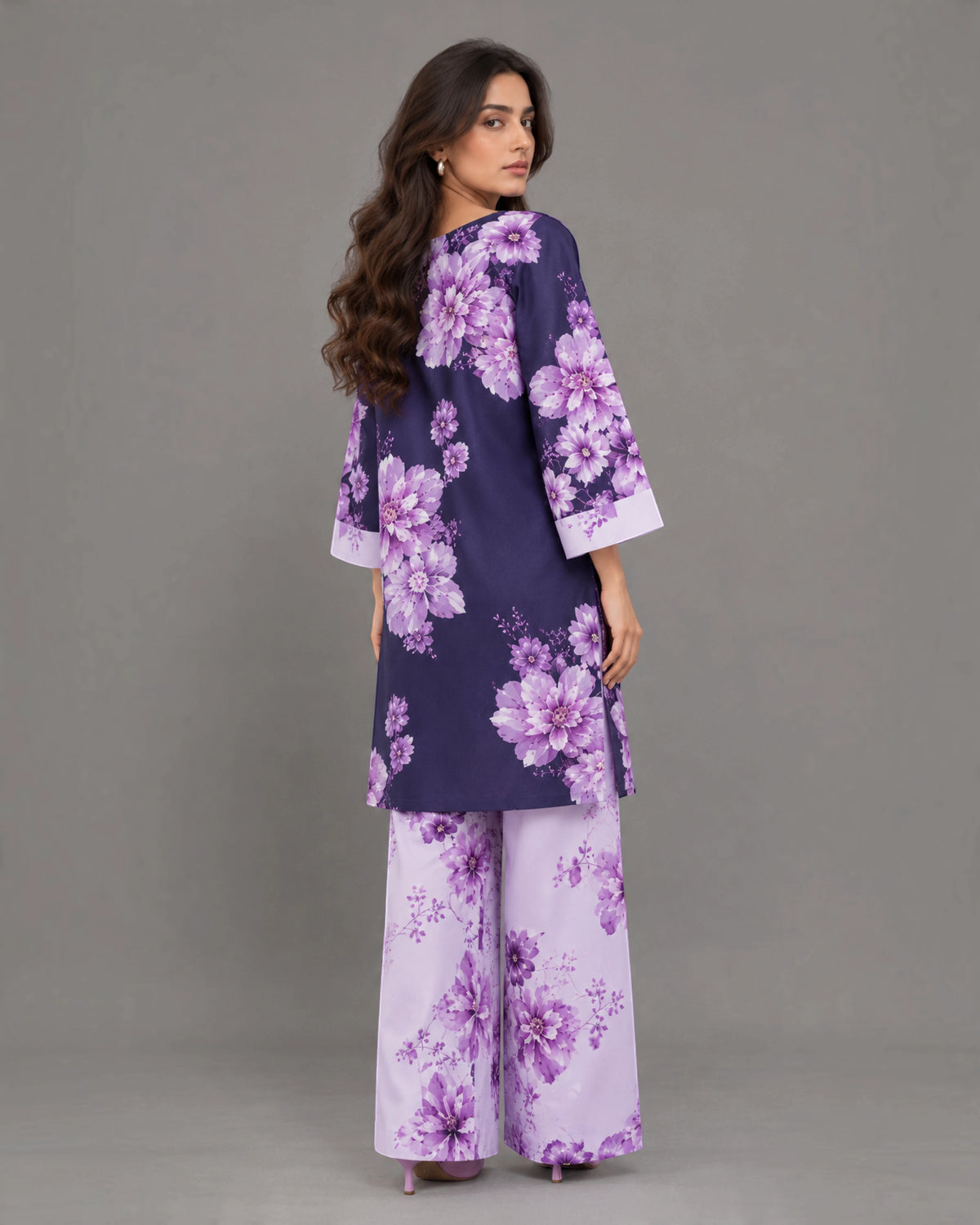 A woman modeling a matching two-piece floral outfit, featuring a navy blue kurta with large purple floral prints and wide-leg trousers in a light lavender base with purple floral patterns, shot against a solid gray studio background.