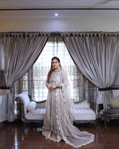 Woman in a beige traditional outfit standing in a decorated room with curtains and furniture.