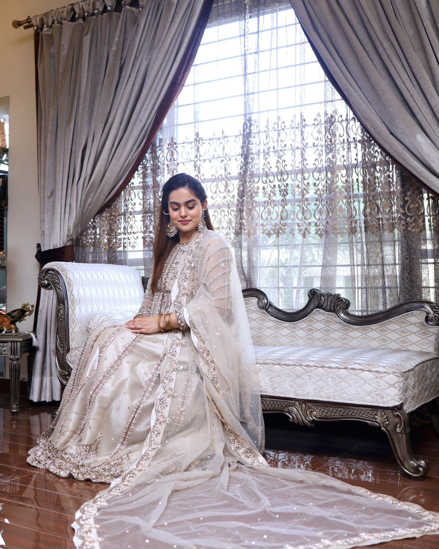 Woman in a white saree sitting on a decorative chair with large windows in the background