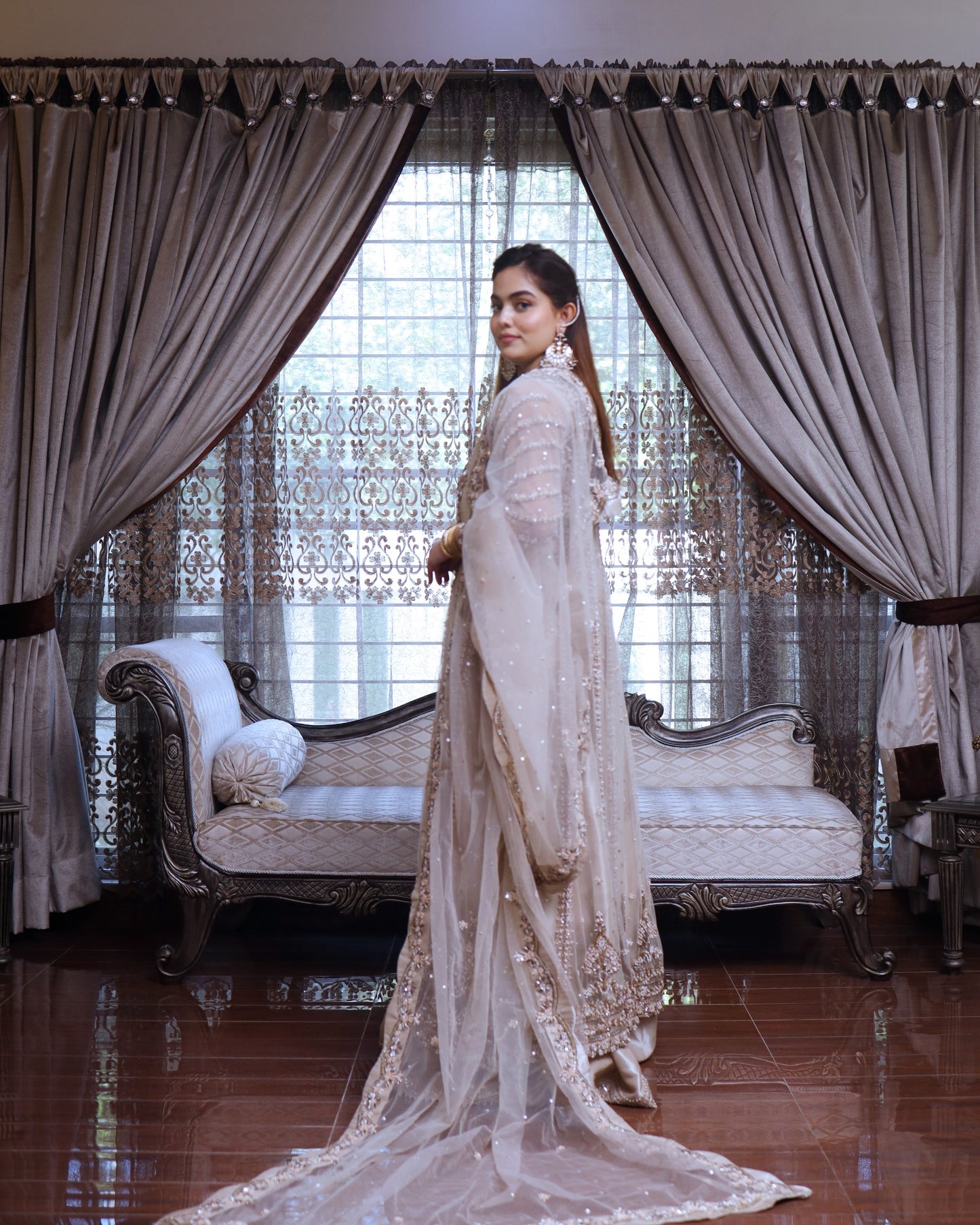 Woman in a white embroidered outfit standing in a decorated room with curtains and furniture.