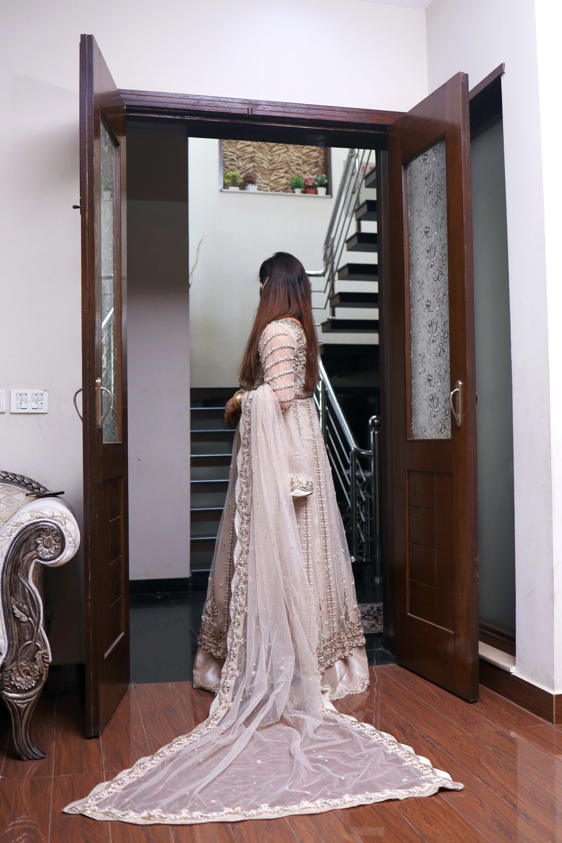 Woman in a beige saree standing in a doorway with a staircase in the background
