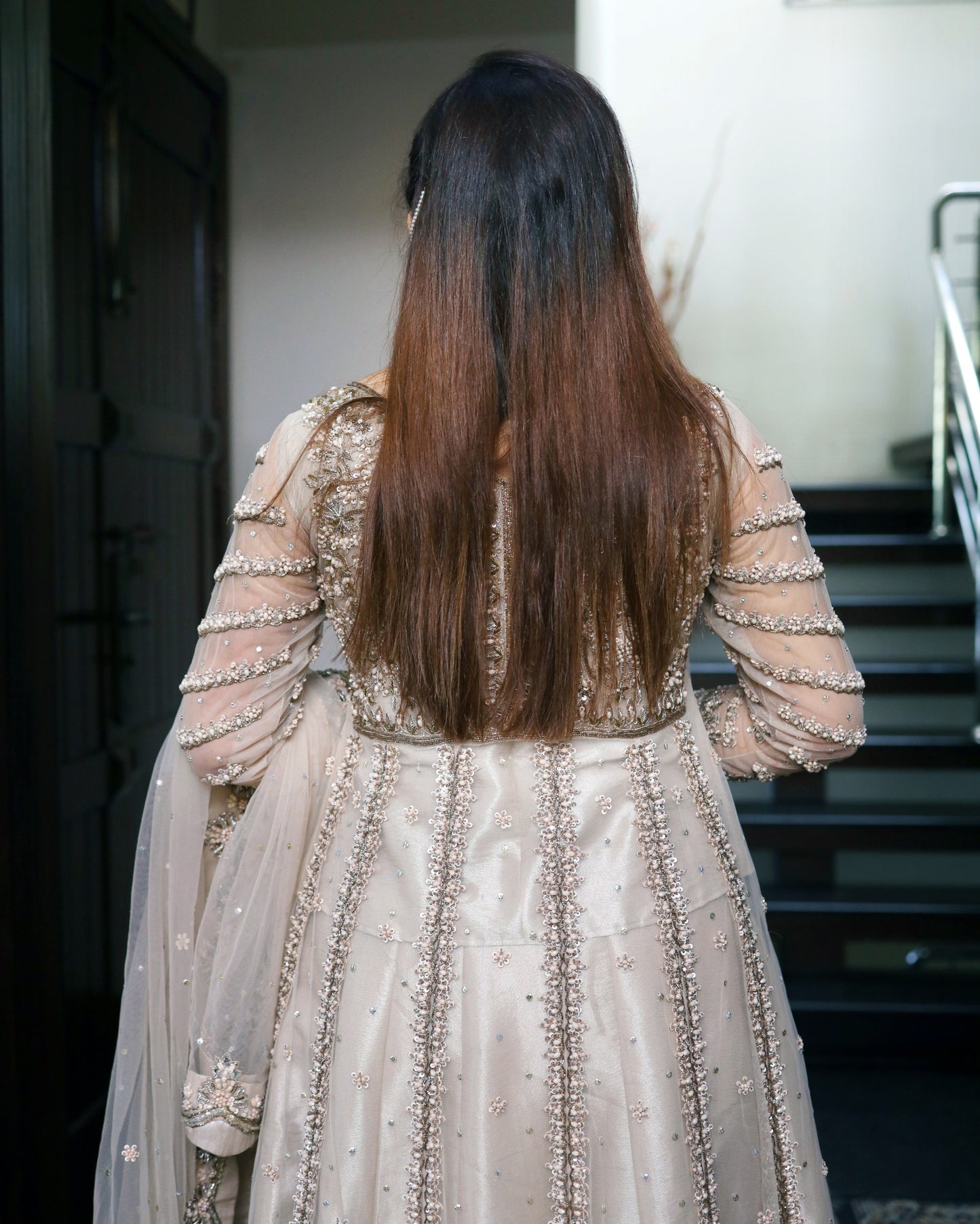 Person wearing a beige embroidered traditional outfit with long hair, standing in a staircase.