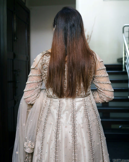 Person wearing a beige embroidered traditional outfit with long hair, standing in a staircase.