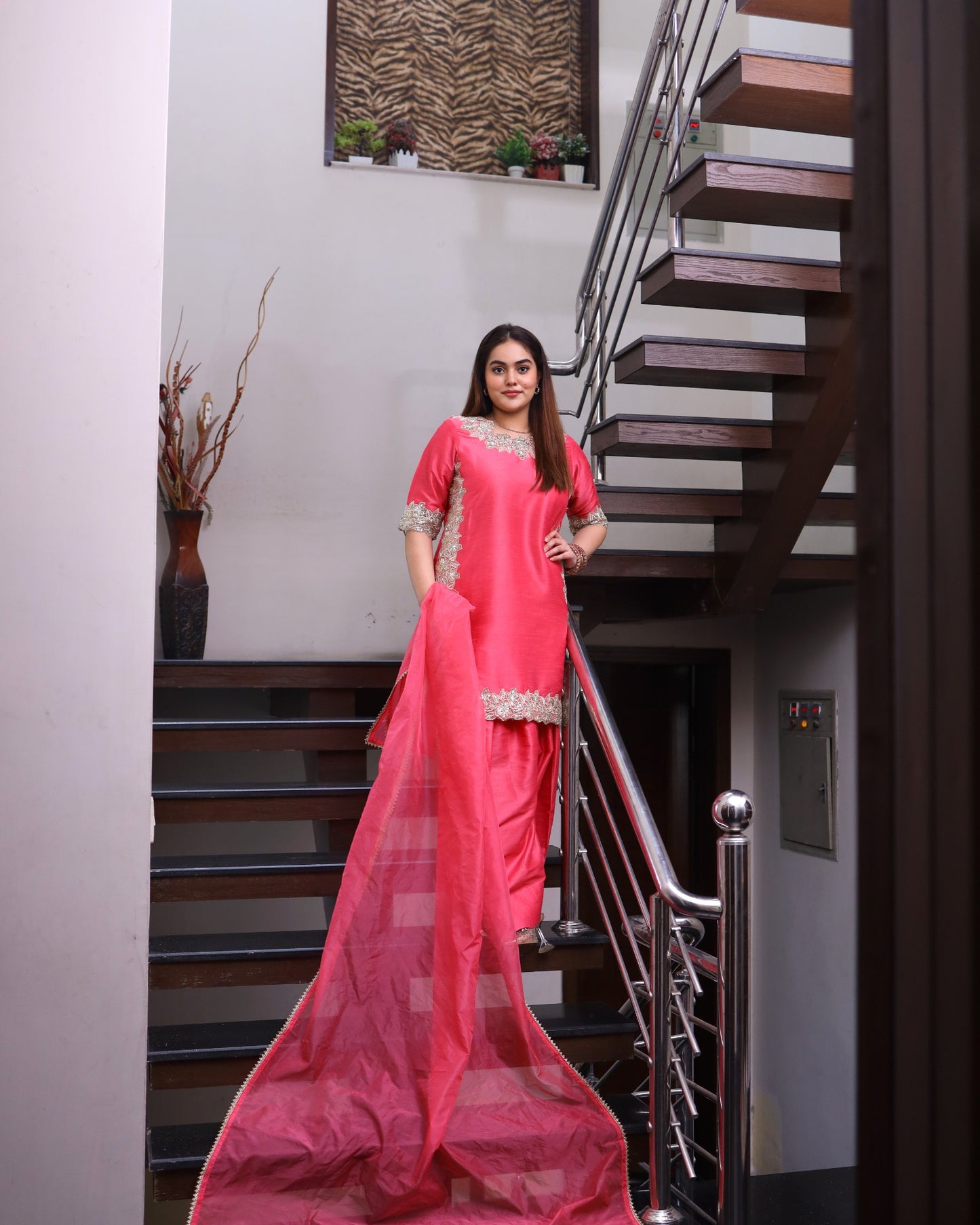 Woman in a pink saree standing in a modern interior setting with stairs.