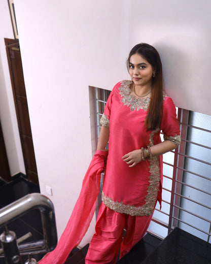 Woman in a pink traditional outfit with gold embroidery standing indoors.