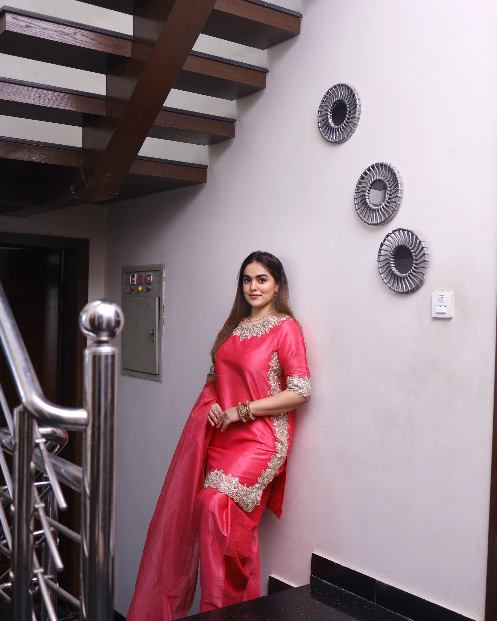 Woman in a pink saree standing in a modern interior setting with decorative wall art.
