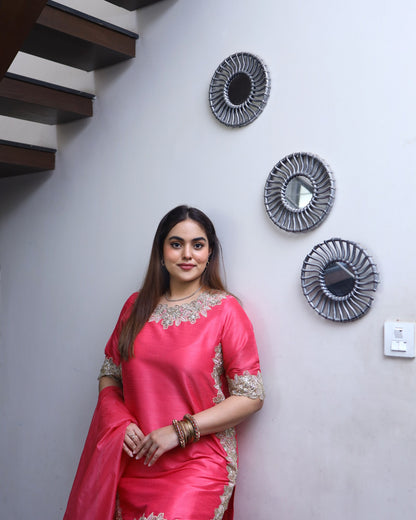 Woman in a pink saree standing against a wall with decorative mirrors.