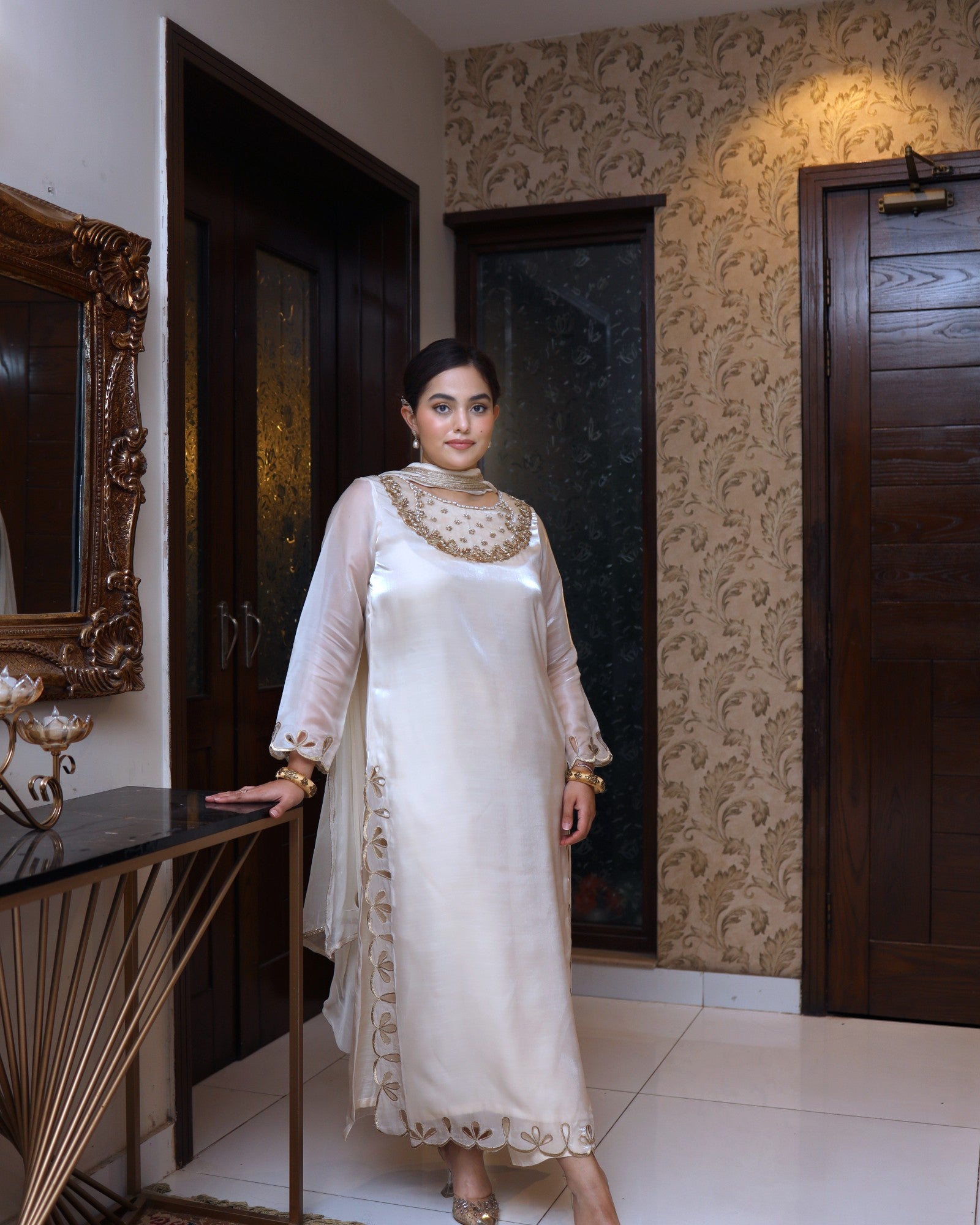 Woman in a white traditional outfit standing in a room with decorative walls and furniture.