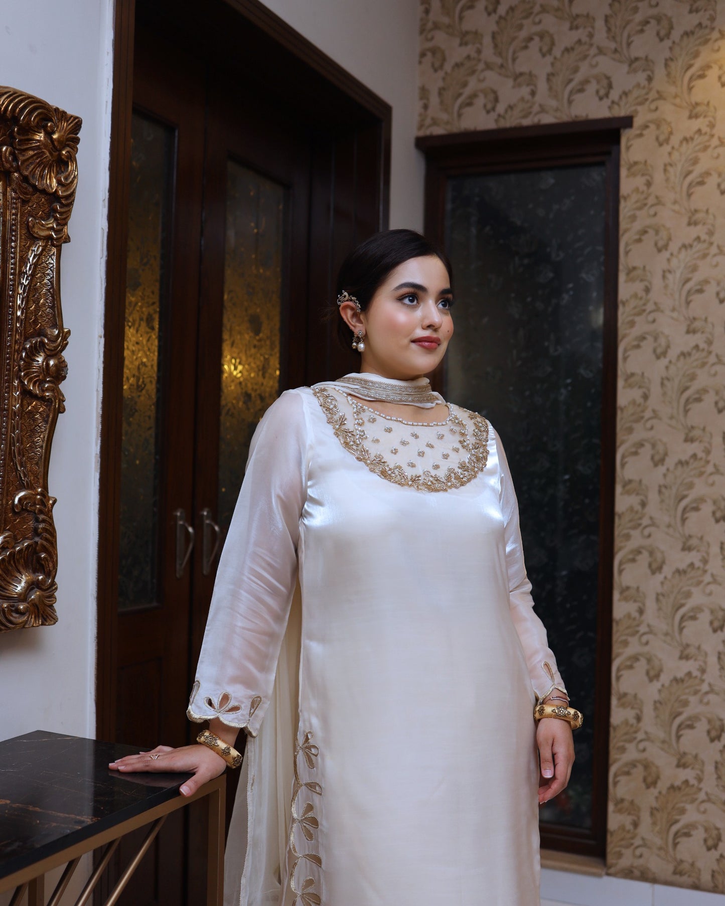 Woman in a white traditional outfit with gold embroidery standing in a room with decorative wall patterns.
