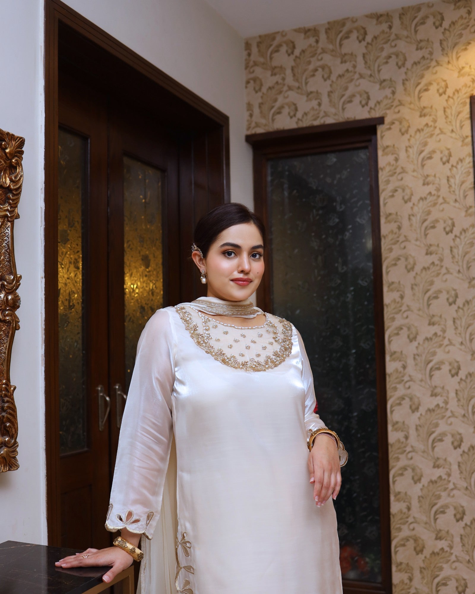 Woman in a white traditional outfit with gold embroidery standing in a room with patterned wallpaper.