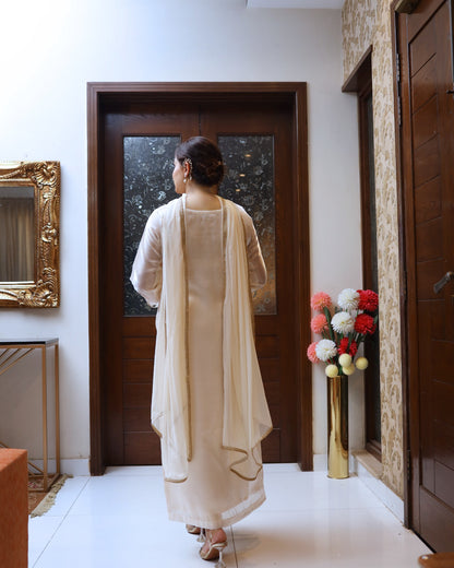 Woman in a beige outfit standing in a room with a decorative mirror and floral arrangement.