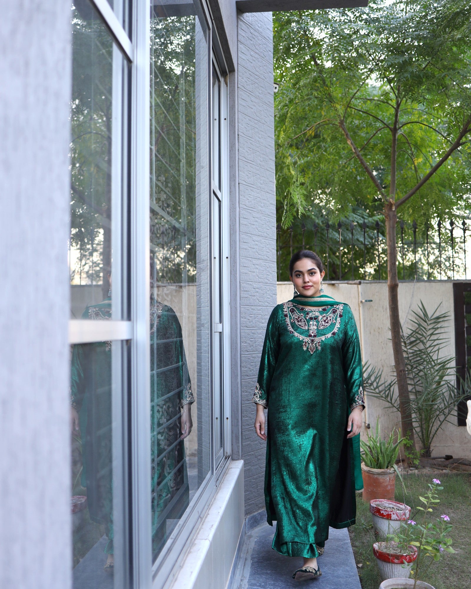 Woman in a green traditional outfit standing by a window with a garden view.