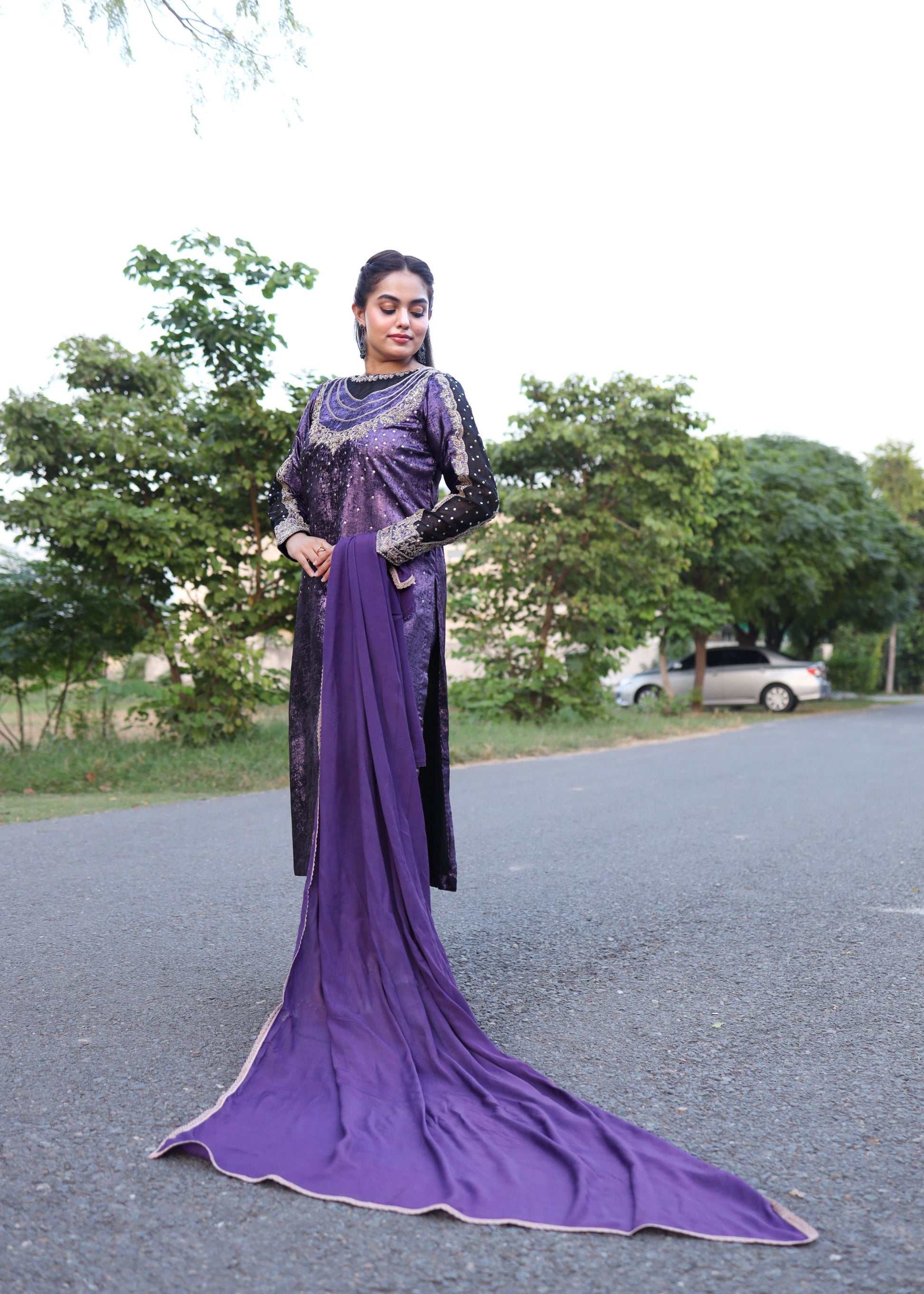 Woman wearing a purple and black traditional outfit standing on a road with trees in the background