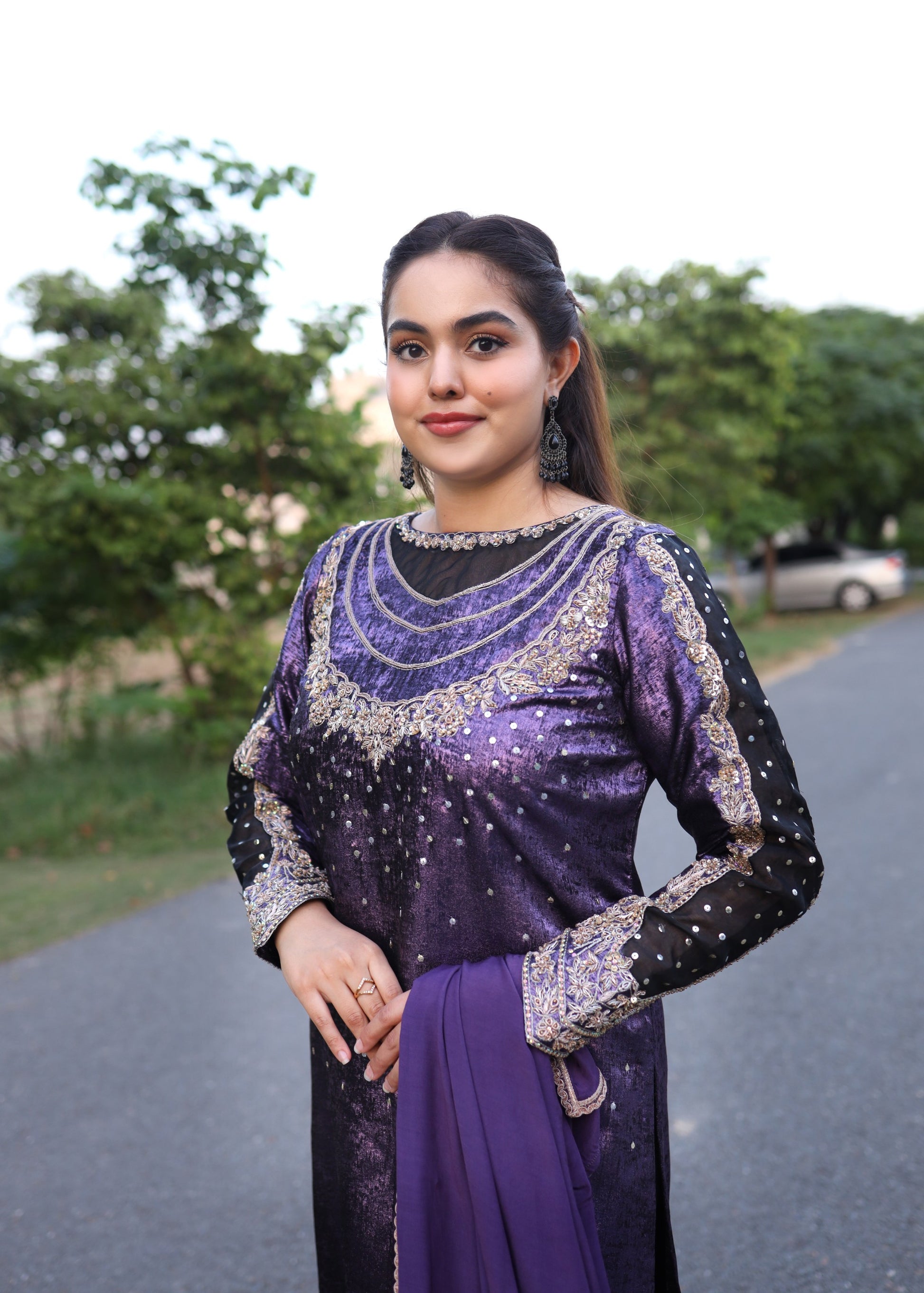 Woman in a purple and black traditional outfit standing outdoors with trees in the background