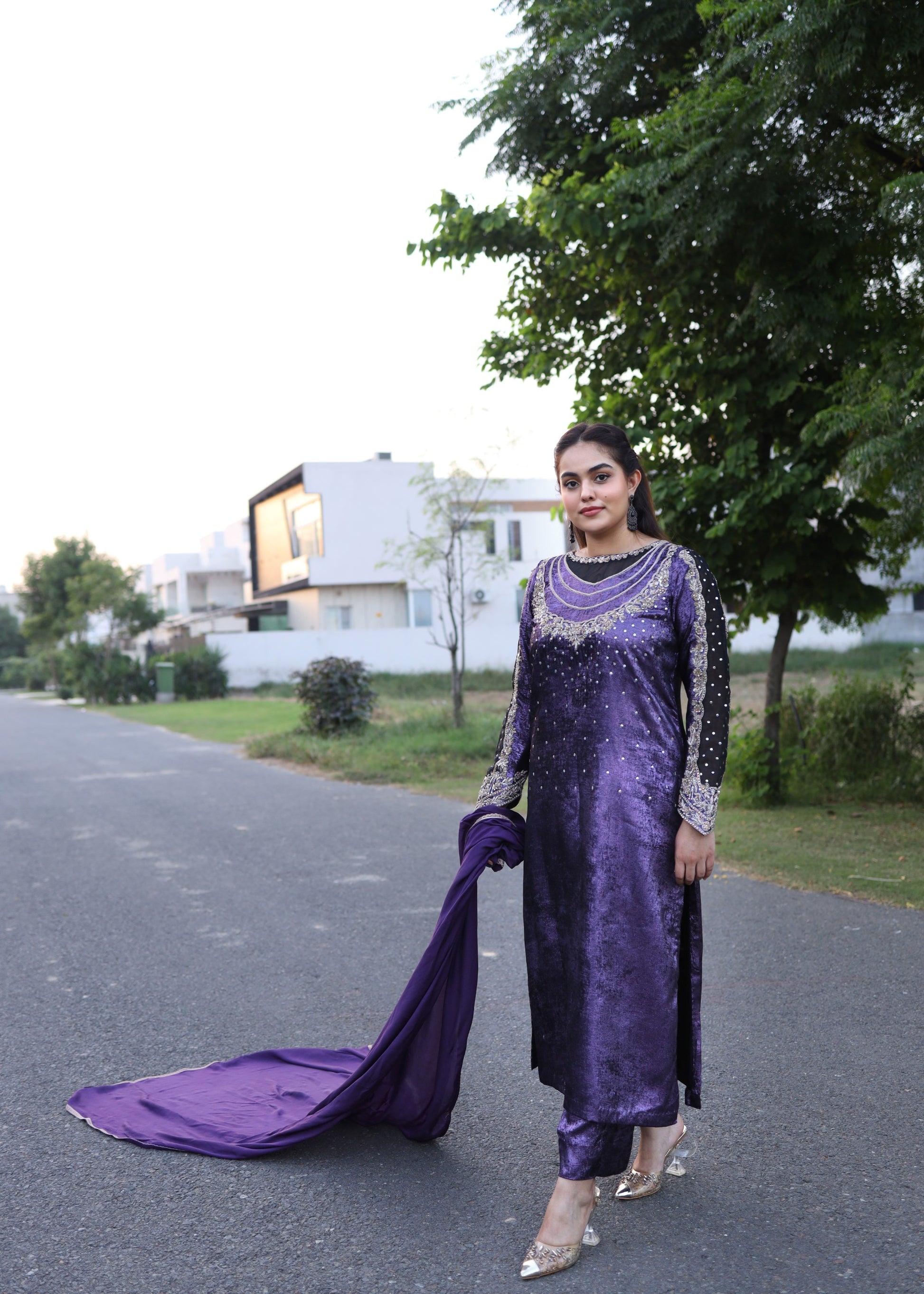 Woman in a purple traditional outfit standing on a road with trees and houses in the background