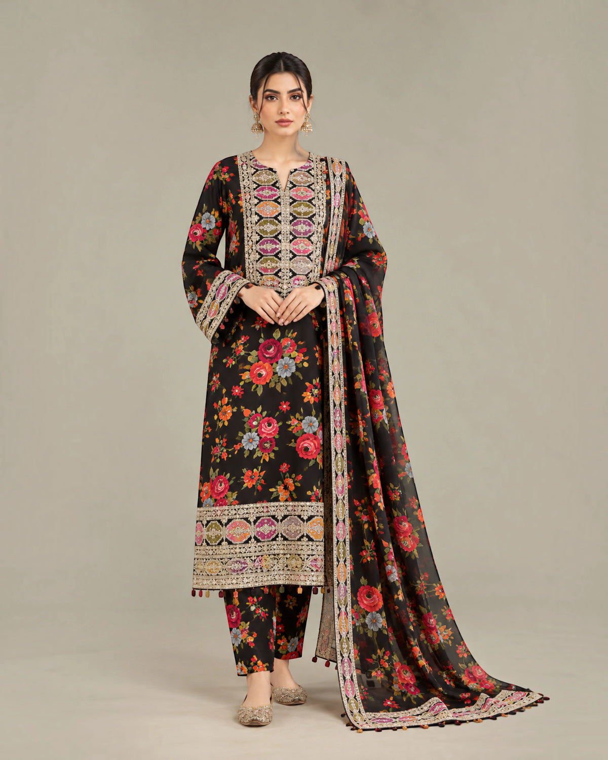 A woman models a traditional three-piece salwar kameez suit in black, featuring a vibrant multicolor floral print, ornate geometric embroidery on the neckline and hem, and a matching printed dupatta, set against a plain beige studio background.