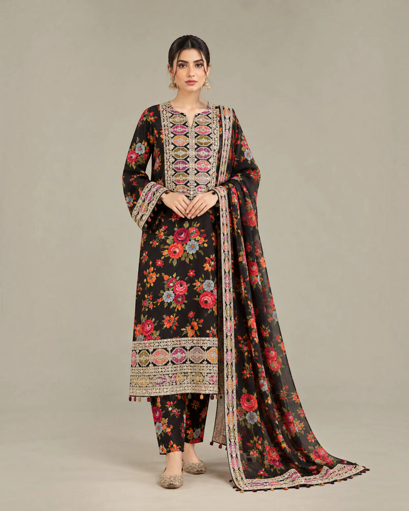 A woman models a traditional three-piece salwar kameez suit in black, featuring a vibrant multicolor floral print, ornate geometric embroidery on the neckline and hem, and a matching printed dupatta, set against a plain beige studio background.