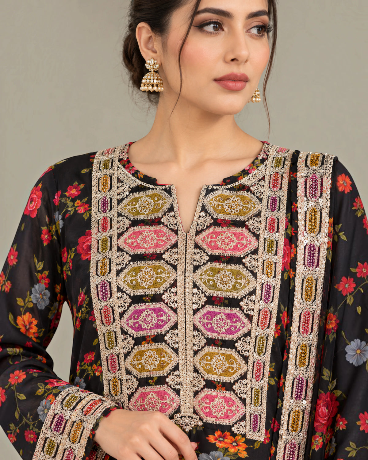 A woman wearing a black floral printed kurta with ornate multicolor embroidery and beadwork on the front panel and cuffs, paired with gold jhumka earrings, against a plain light gray background.