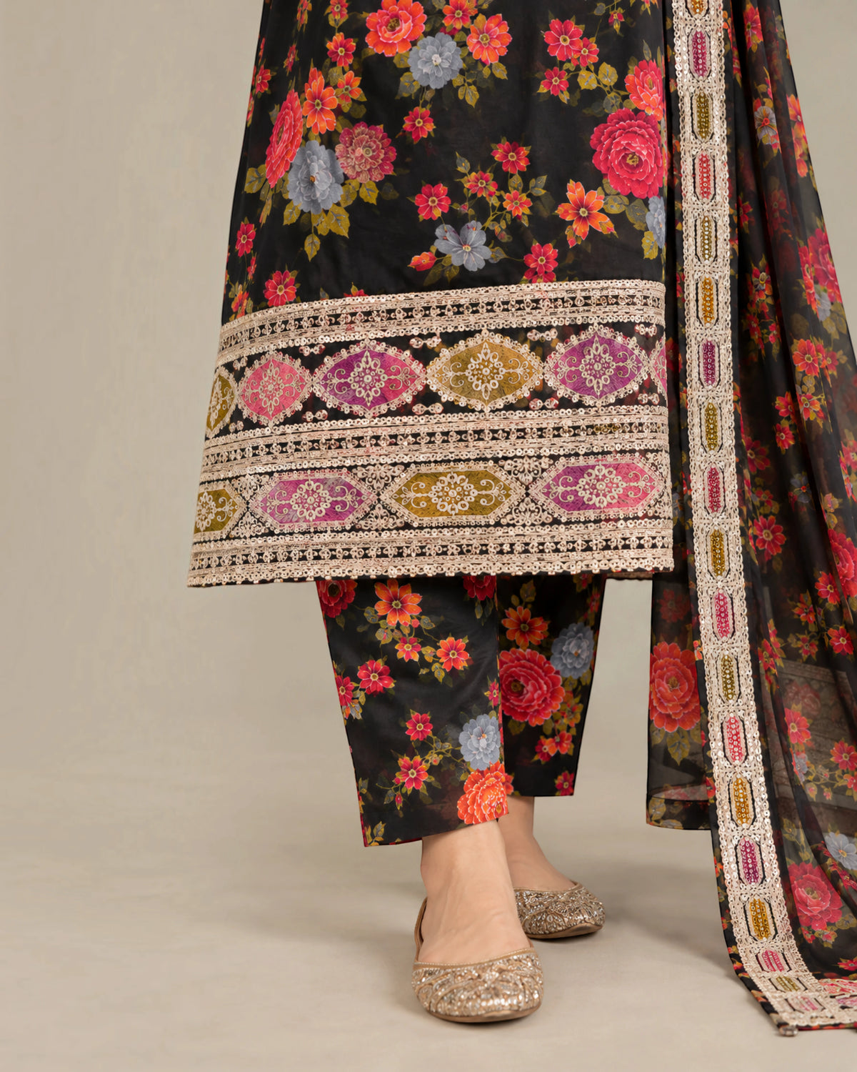 A close-up of a model wearing a black three-piece ethnic suit with a vibrant floral print, featuring detailed multicolor embroidery on the hem and a matching dupatta, paired with glittery flat shoes.