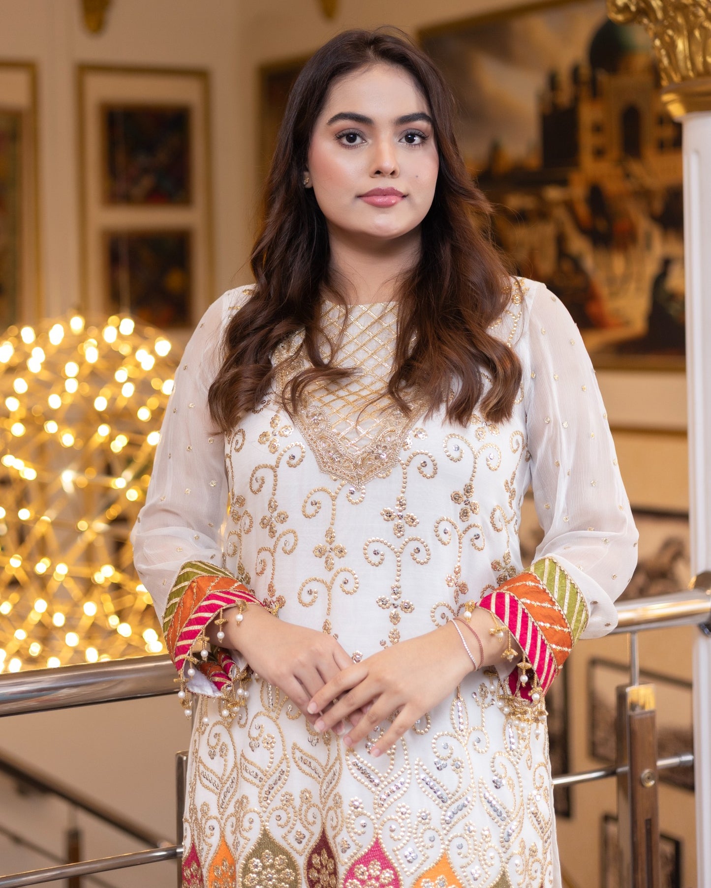 Woman in an embroidered white dress with colorful bangles standing indoors.