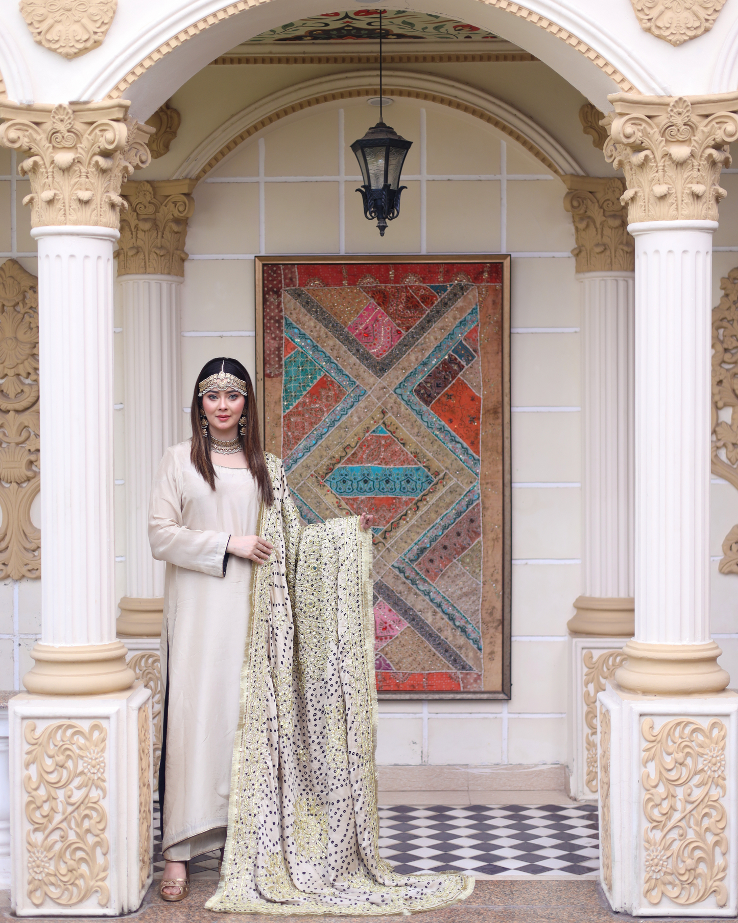 Woman in a white dress with a patterned scarf standing in an ornate architectural setting.
