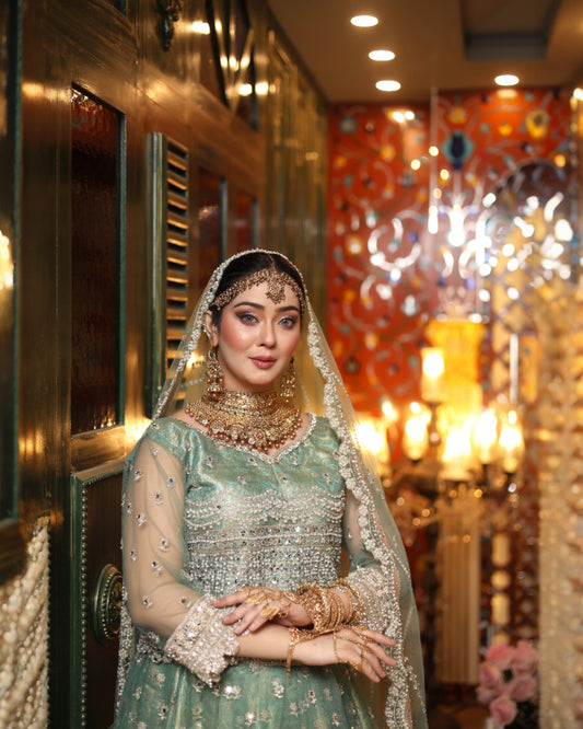 Woman in traditional green bridal outfit with jewelry in a decorated room.