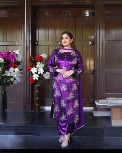 Woman in a purple traditional outfit standing in front of a wooden panel wall with floral decorations.