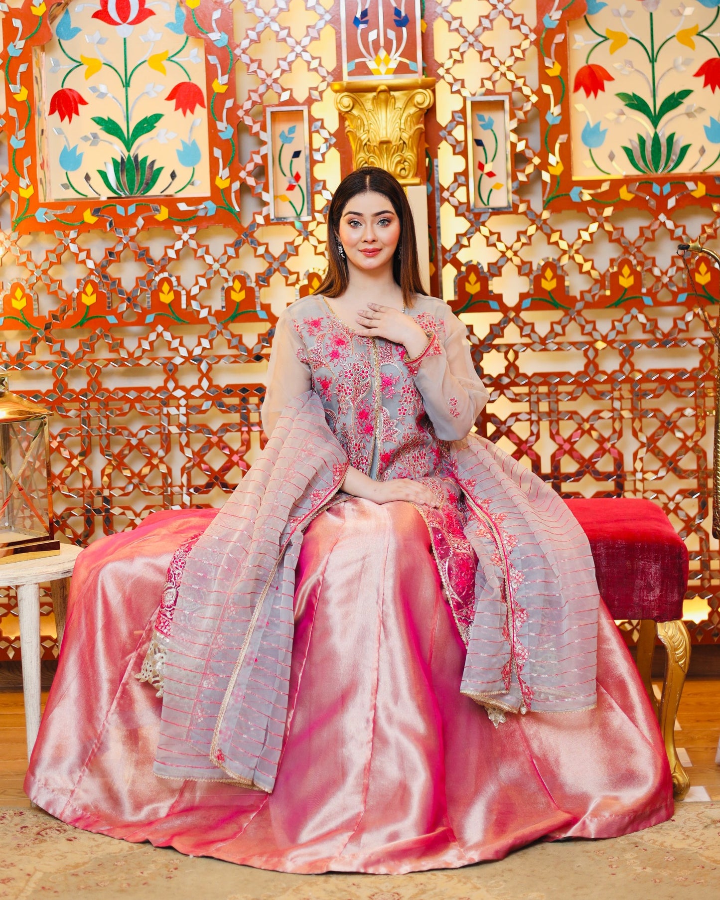 Woman in a pink and white embroidered traditional outfit sitting on a decorative chair with a colorful patterned wall behind.