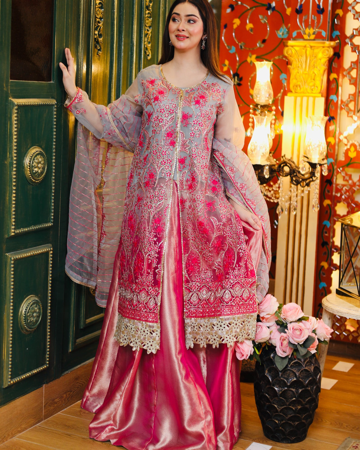 Woman in a pink and gray embroidered traditional outfit standing in a decorated room.