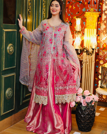 Woman in a pink and gray embroidered traditional outfit standing in a decorated room.