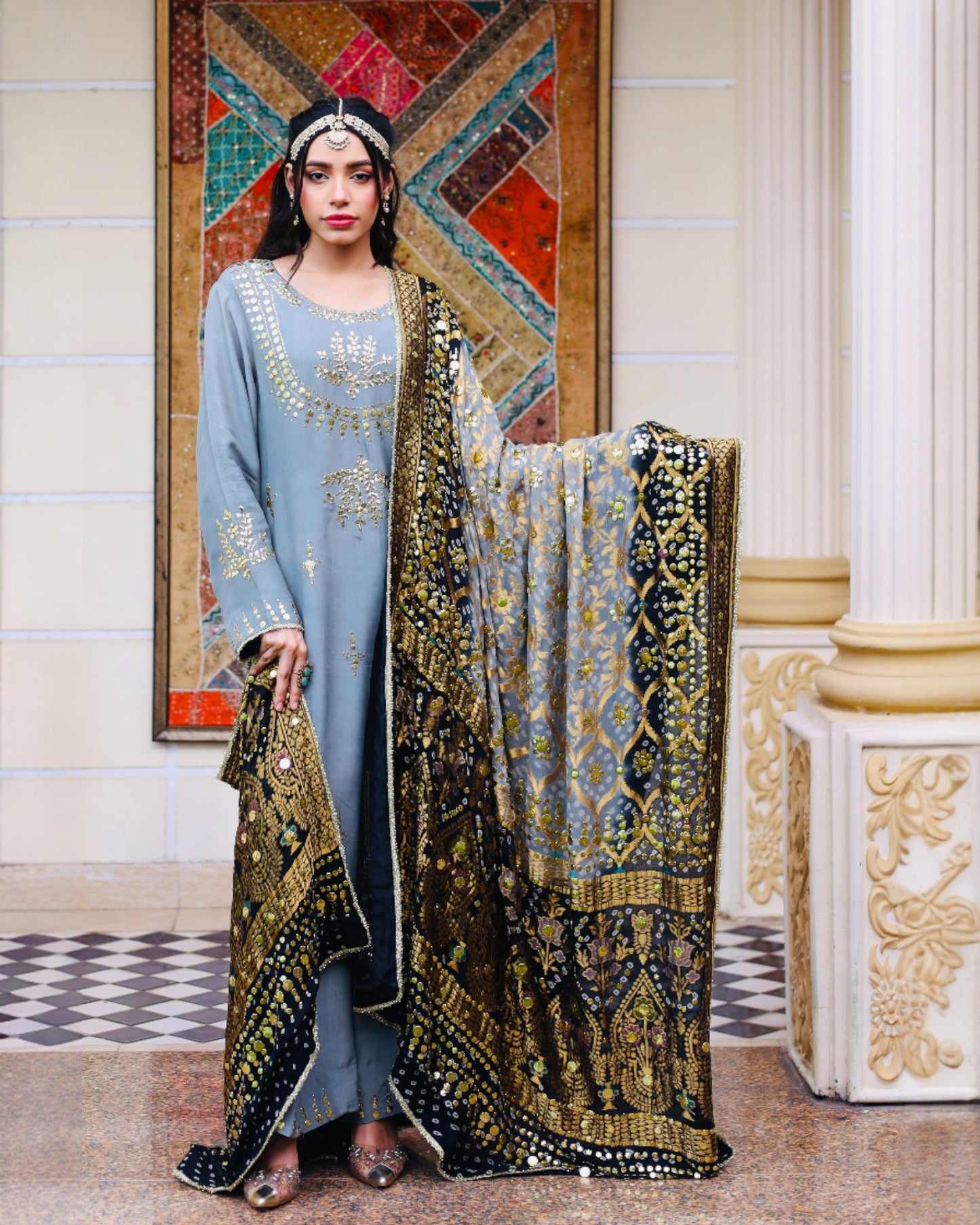 Woman in a blue traditional outfit with a patterned scarf in an ornate room.