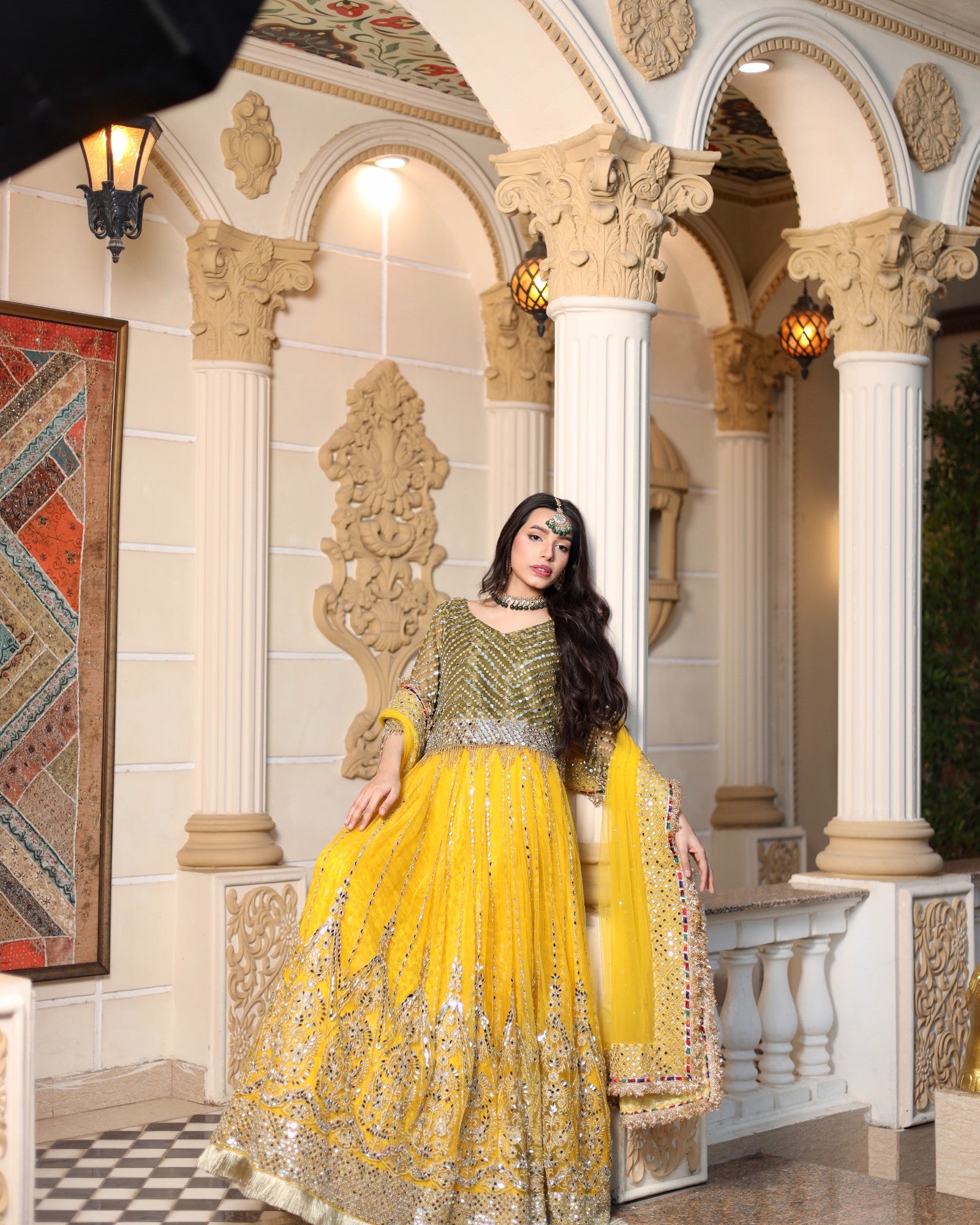 Woman in a traditional yellow and green outfit sitting in an ornate architectural setting.