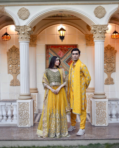 Two individuals in traditional yellow and green outfits standing in an ornate architectural setting.