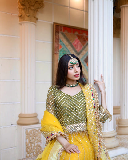 Woman in traditional yellow and green outfit with jewelry, standing indoors.