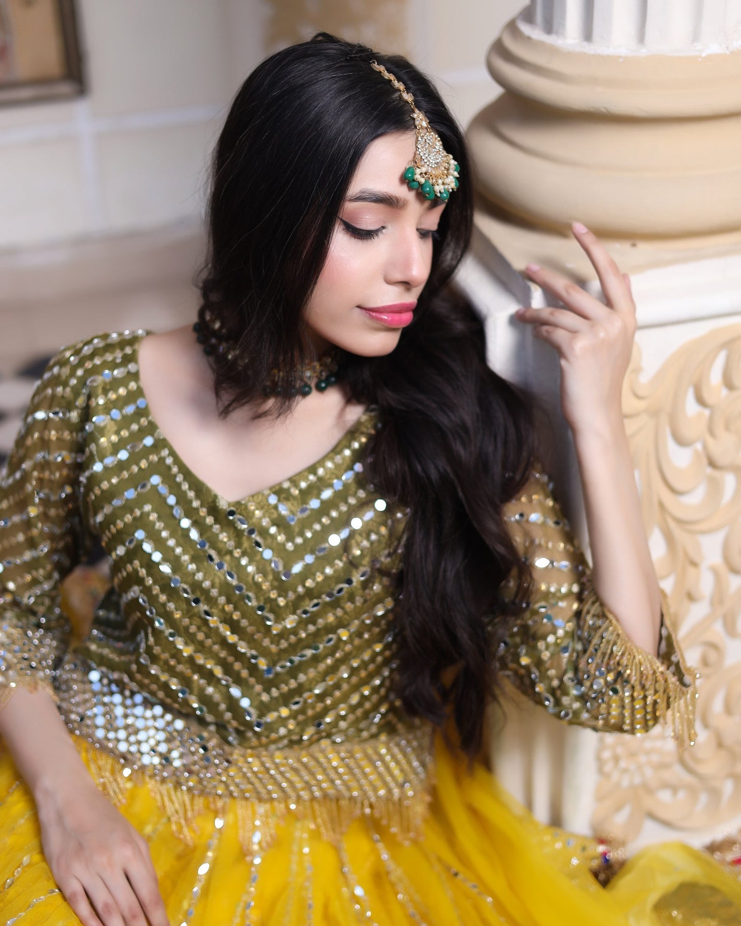 Woman in a gold and yellow traditional outfit with jewelry, sitting indoors.