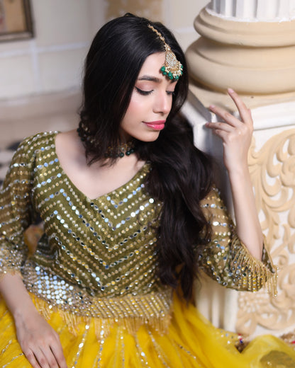Woman in a gold and yellow traditional outfit with jewelry, sitting indoors.