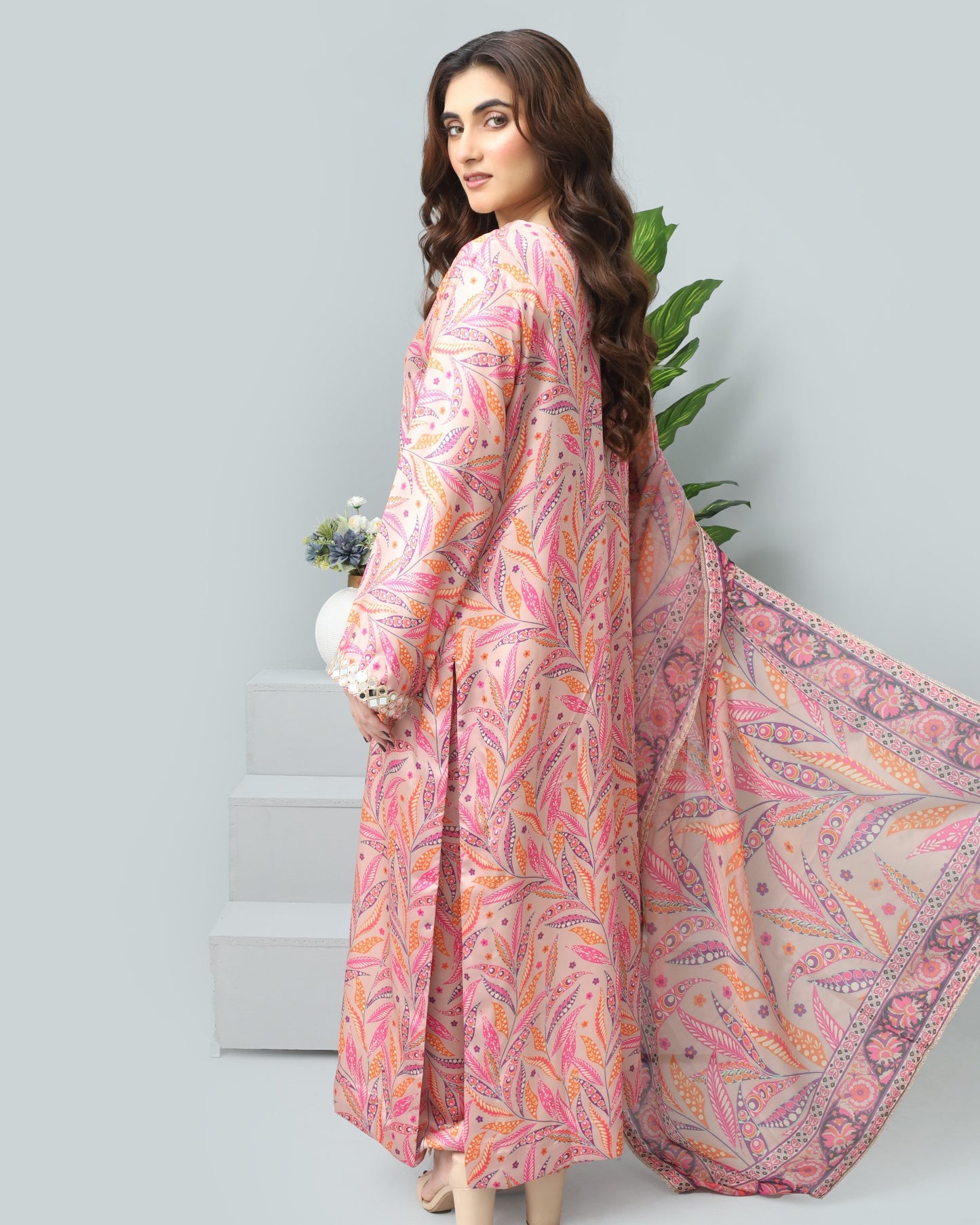 Woman wearing a pink and orange patterned dress with a scarf, standing against a light gray background.