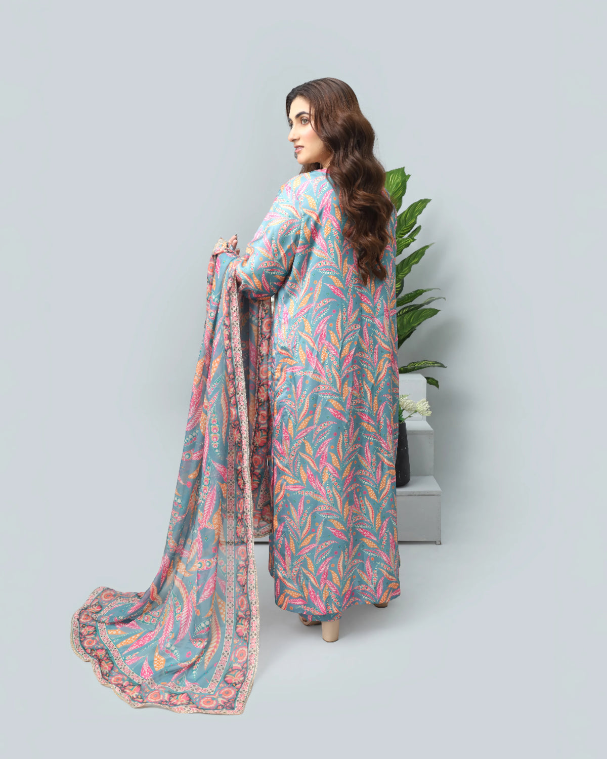 A woman stands in profile wearing a long-sleeved, loose-fitting tunic with a colorful abstract leaf-like print in pink, orange, and blue on a light gray base, holding a matching patterned dupatta. She is photographed against a plain light gray studio background with a small potted plant behind her.