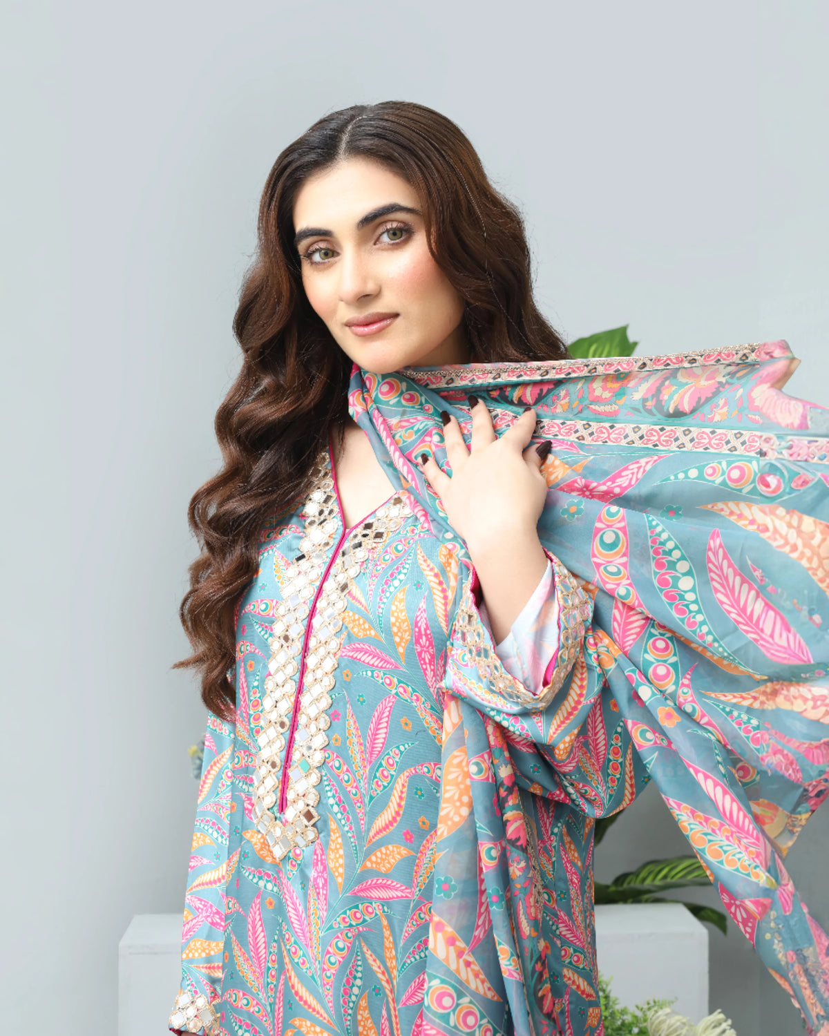 A woman modeling a light blue printed traditional outfit with pink, orange, and green patterns, holding a matching dupatta against a light gray background.