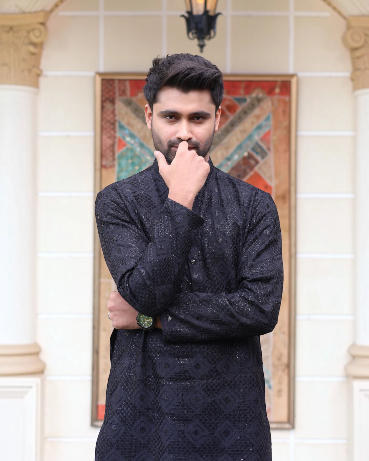 A man wearing a black textured kurta stands in front of a cream-colored stone wall with columns and a decorative panel.
